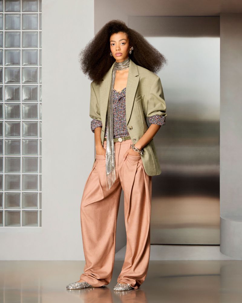 Model with voluminous hair wearing a sage green blazer, metallic mesh scarf, and wide-leg trousers in a modern studio setting.