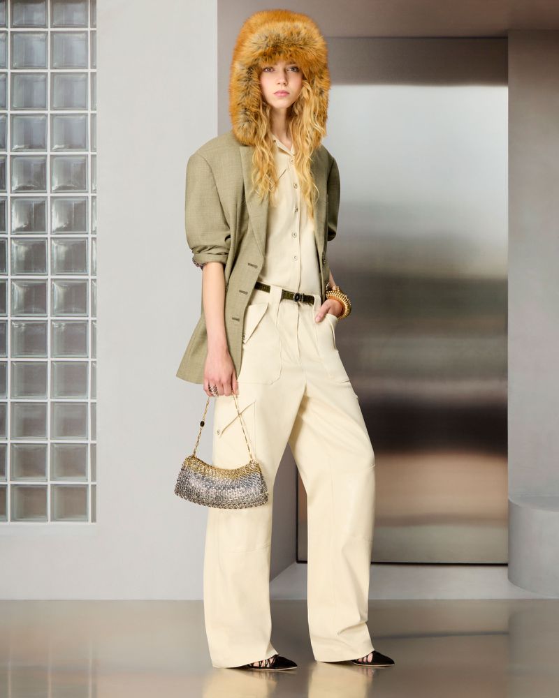 Model wearing a large amber fur hat, olive blazer, cream trousers, and holding a metallic chainmail bag against a glass block wall.