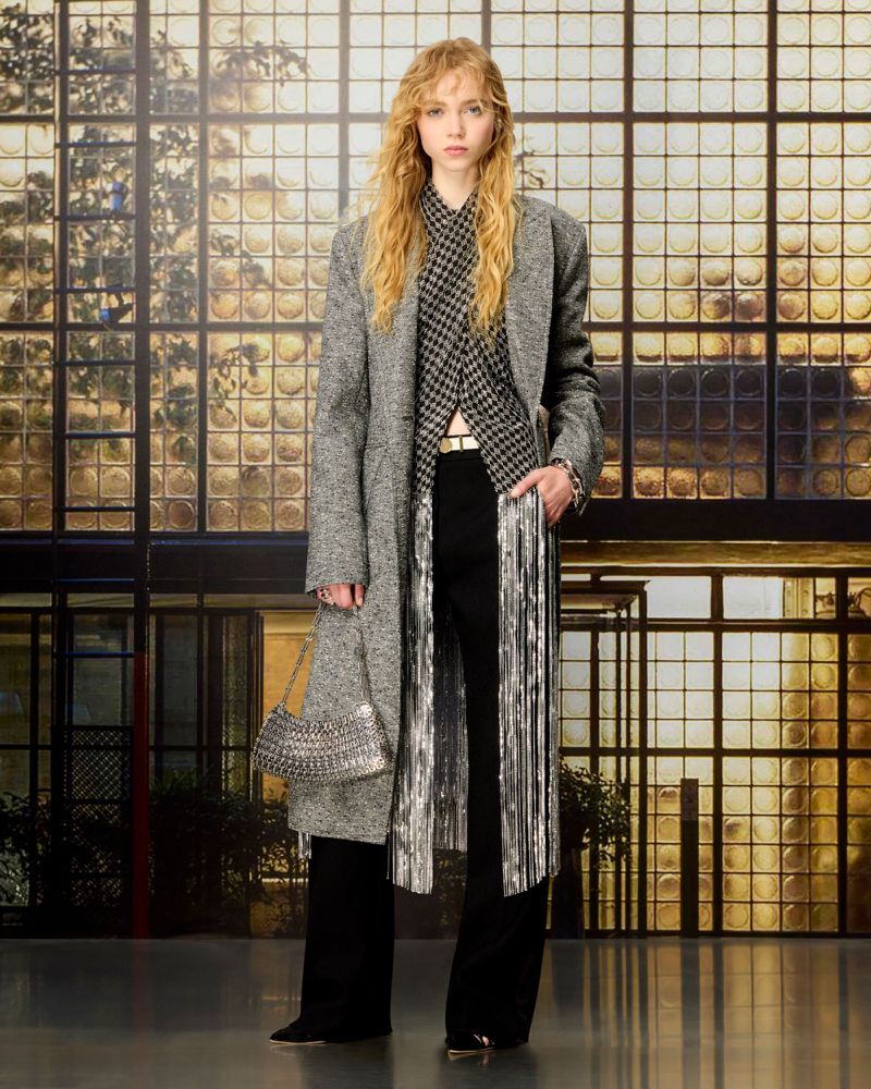 Model standing in a long grey tweed coat, houndstooth top, metallic fringe, and black trousers holding a silver mesh bag.