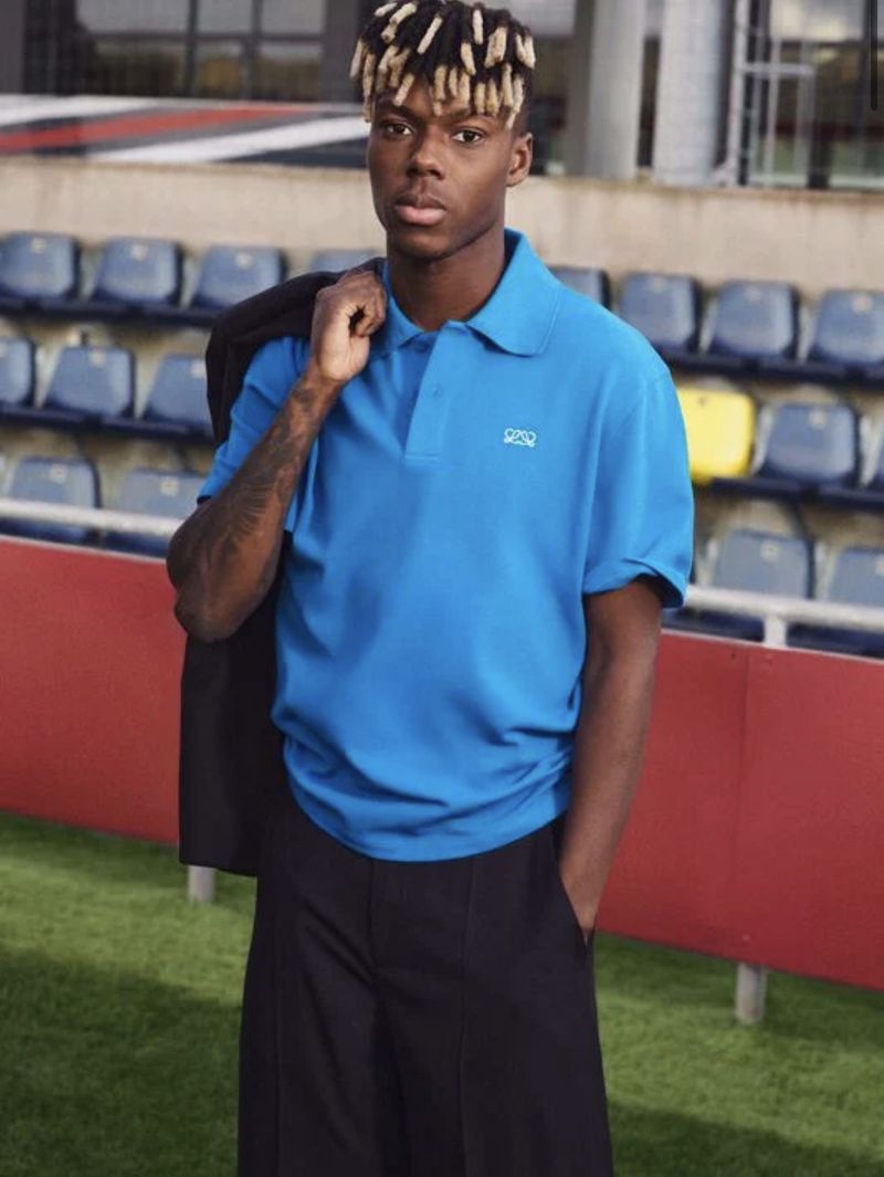 Spanish footballer Nico Williams wearing a bright blue Loewe polo shirt and black trousers in a sports stadium.