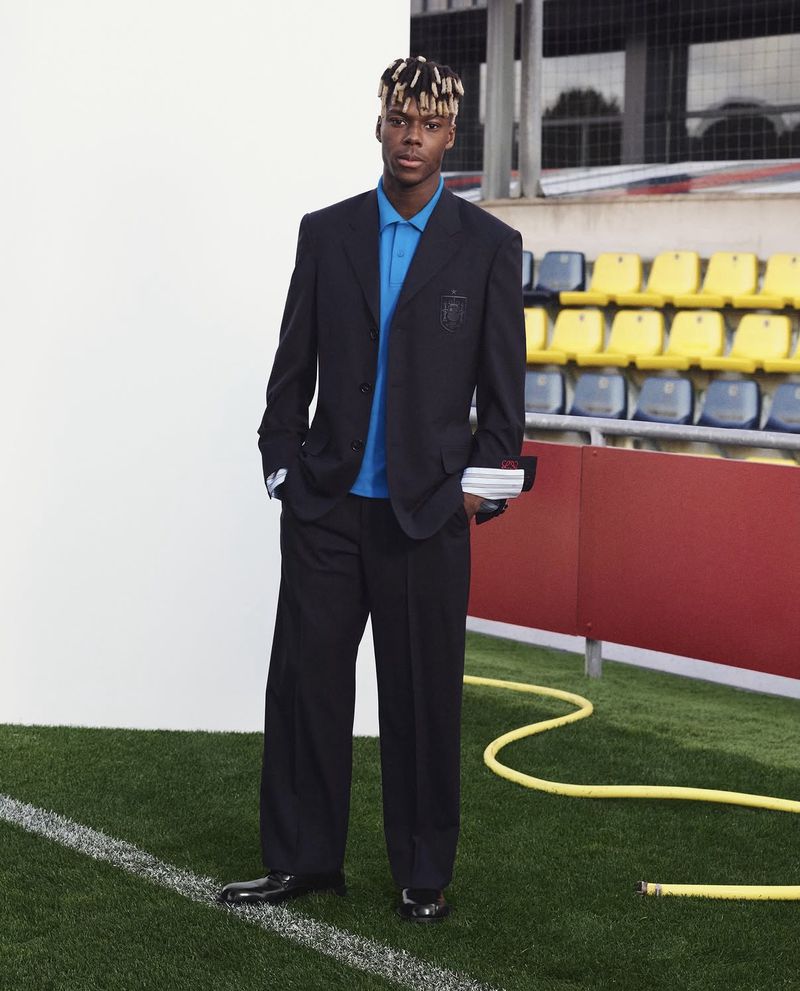 Footballer Nico Williams posing in a black Loewe tailored suit and bright blue polo shirt pitchside at a stadium.