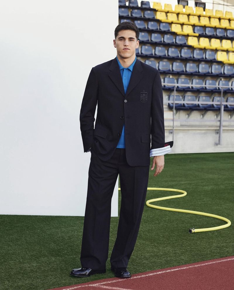Footballer Pau Cubarsí stands in a dark navy tailored suit and bright blue polo shirt by a white backdrop on a stadium pitch.