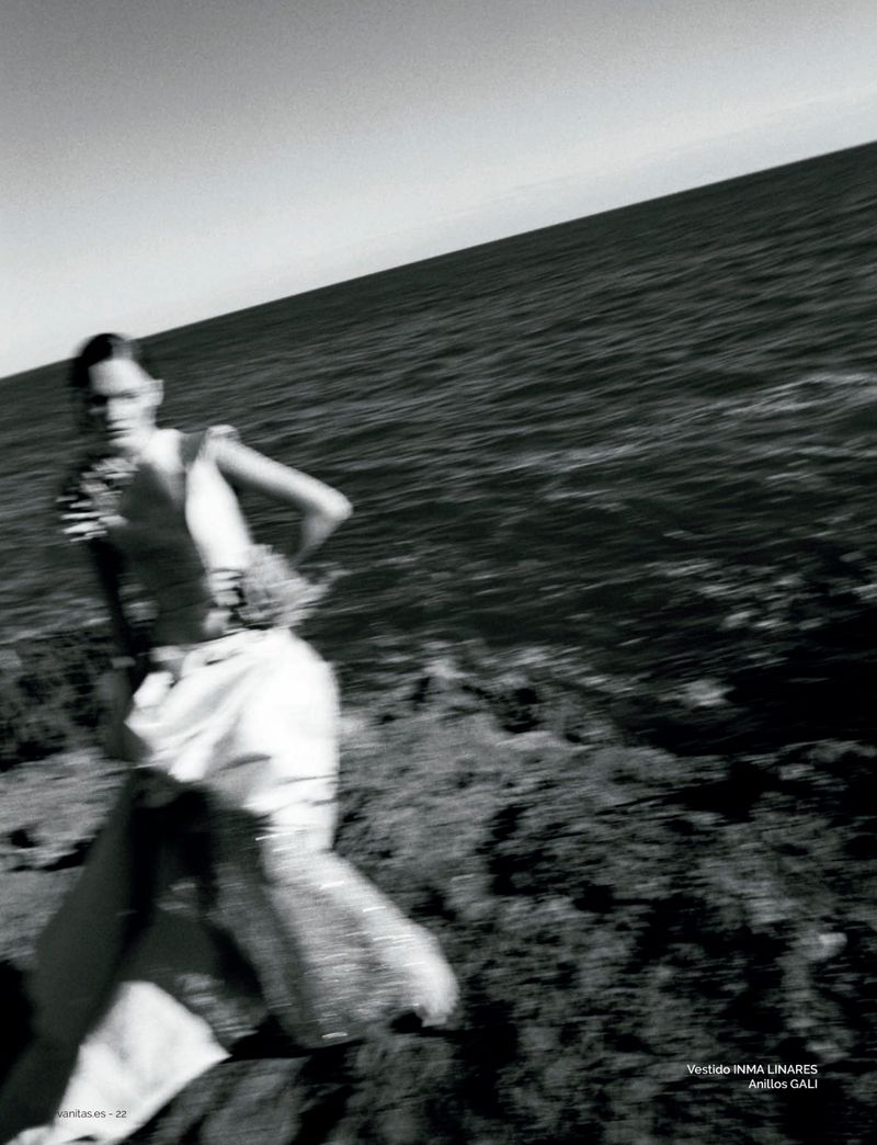 Model wearing a light dress moving across a rocky coast with the ocean behind in a black and white editorial shot.