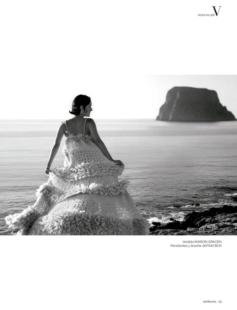 Black and white editorial photograph of a woman in a voluminous tiered dress standing on a rocky shore looking out at the sea.