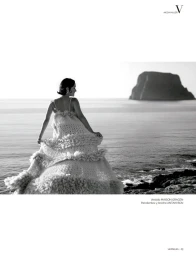 Black and white editorial photograph of a woman in a voluminous tiered dress standing on a rocky shore looking out at the sea.