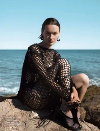 Model seated on coastal rocks wearing a black sequined open-knit dress and mesh heels against the blue sea.