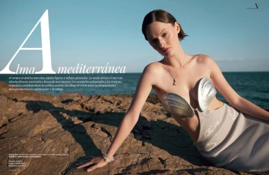 Model reclining on a rocky coast wearing a metallic clam shell bra top and light grey skirt beside the ocean.