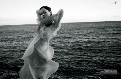 Woman in a sheer sculptural ruffled dress posing against the open sea in a black and white editorial shoot.
