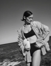 Black and white photo of a woman in a black bikini, feather-trimmed coat, and seashell clutch walking along a coast.