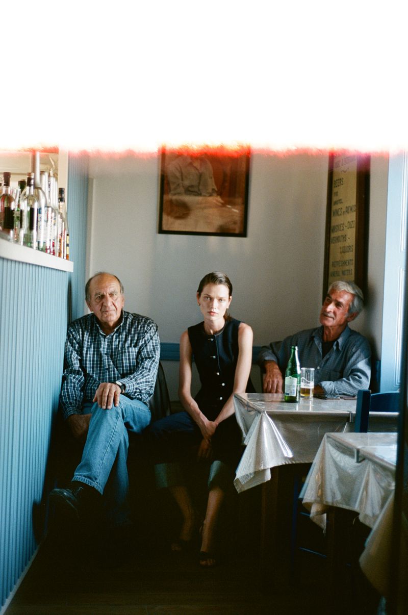 Model Dominika Drozdowska sits between two older men at a small dining table inside a rustic café interior.