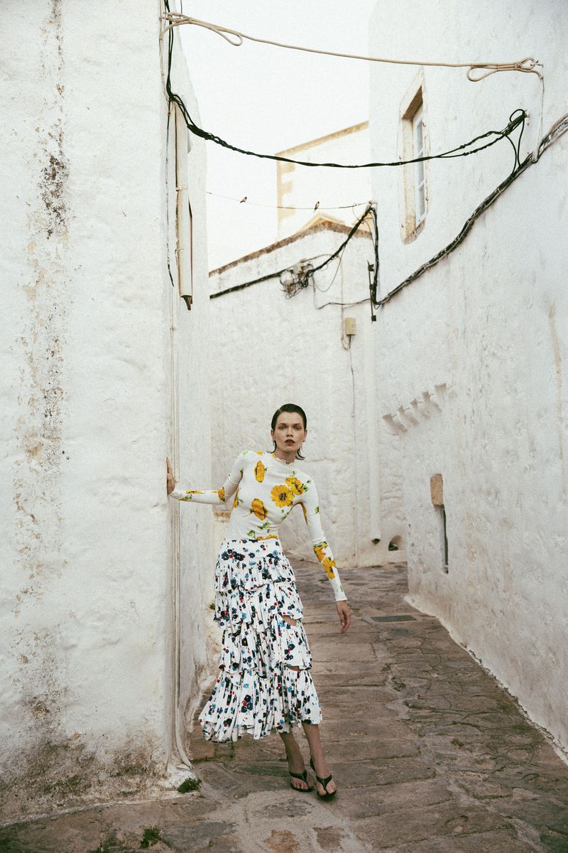 Dominika Drozdowska posing in a yellow floral top and tiered skirt between whitewashed walls in a narrow alley for Elle Greece.