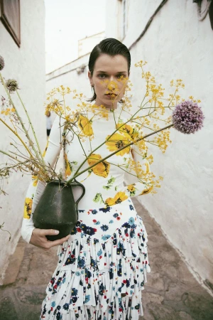 Model Dominika Drozdowska in a floral print top and ruffled skirt holding a dark jug filled with wildflowers in a narrow alley.