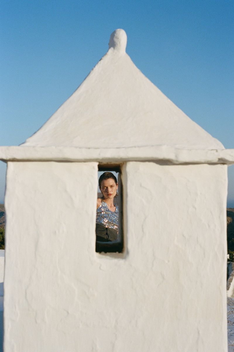 Model Dominika Drozdowska framed through a narrow vertical window of a whitewashed structure under a clear blue sky.
