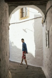 Model Dominika Drozdowska walks through a sunlit whitewashed alley in a blue jacket and floral skirt framed by an archway.