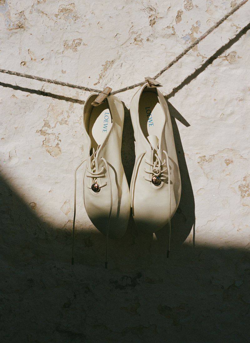Pair of off-white pointed lace-up flat shoes hanging from a clothesline against a weathered plaster wall in direct sunlight.