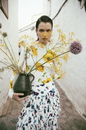 Model Dominika Drozdowska in a floral print top and ruffled skirt holding a dark jug filled with wildflowers in a narrow alley.