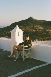 Model Dominika Drozdowska seated in a white director chair on an outdoor terrace with rolling hills in the background.
