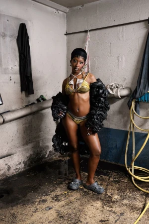 A Black woman posing in a gold bikini, dark ruffled sleeves, and grey clogs in an industrial concrete basement.