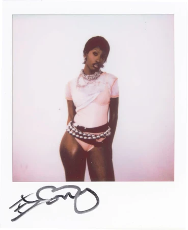 Polaroid photo of a woman with short dark hair in a pink top and studded belt posing against a white background.