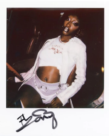 Polaroid photo of Ebony seated inside a car wearing a white cropped top, jacket, and shorts, signed on the bottom border.