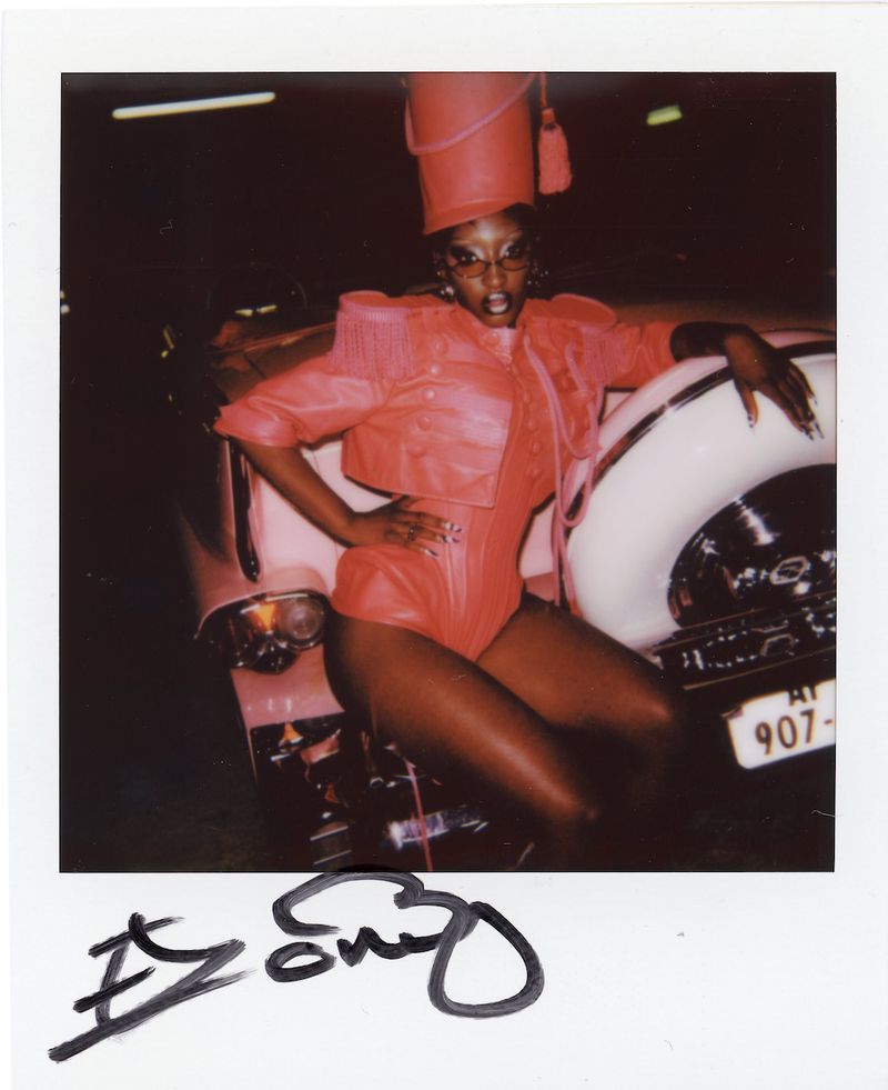 Polaroid photograph of a Black woman in a red marching band uniform posing on a classic car.