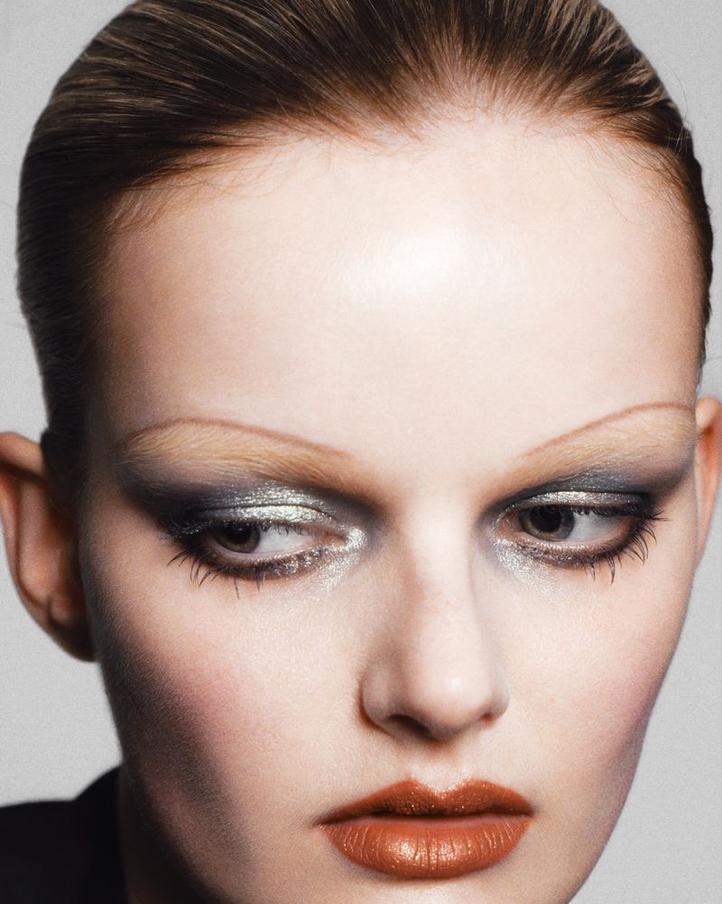 Close-up beauty portrait of a woman with metallic silver eyeshadow, pencil-thin eyebrows, and warm terracotta lipstick.