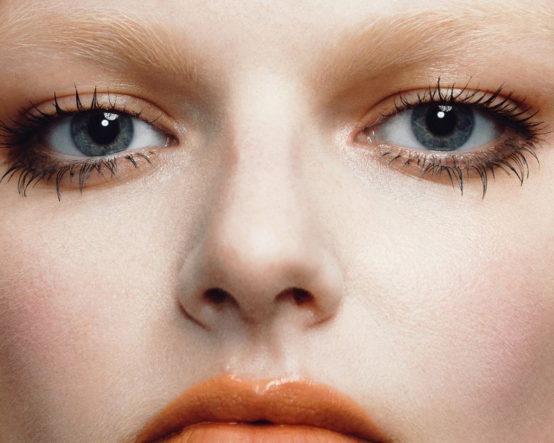 Close-up portrait of a model with blue eyes, bleached brows, and glossy orange lipstick captured by Irina Shestakova.