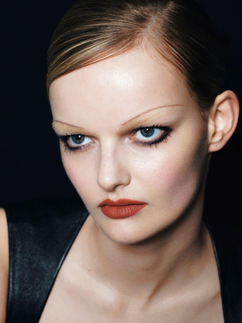 Close-up portrait of a woman with slicked-back hair, striking dark eyeliner, and red lipstick against a dark background