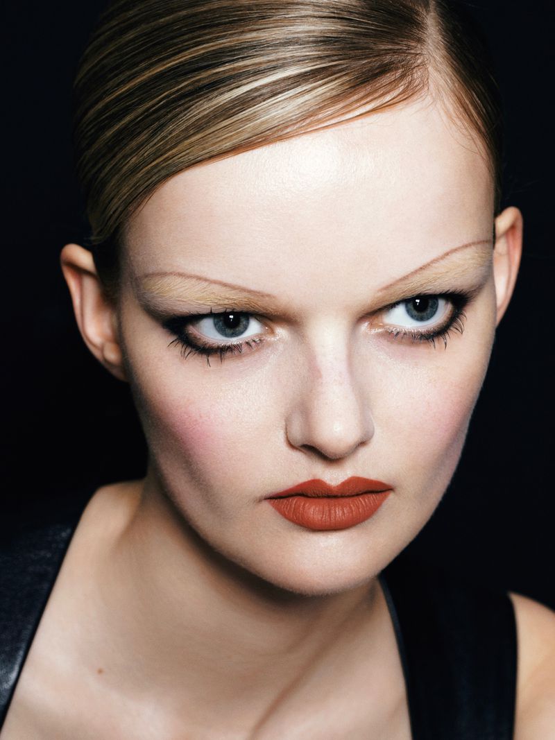 Close-up portrait of a model with sleek blonde hair, bold eyeliner, and dark red lipstick against a black background.