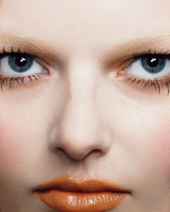 Extreme close-up beauty portrait of a woman with blue eyes, light blonde eyebrows, and glossy orange lipstick.
