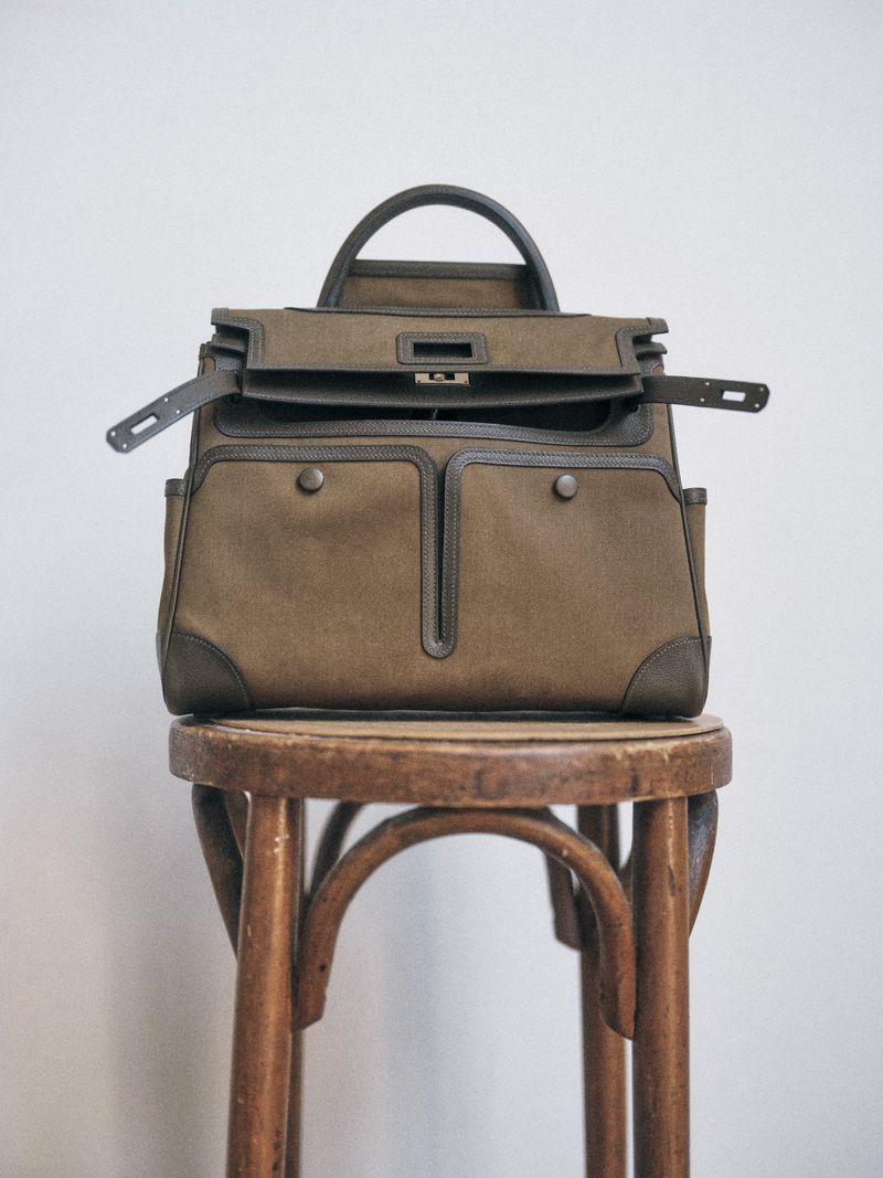 Olive canvas and leather handbag resting open atop a weathered wooden bentwood stool against a plain pale wall.