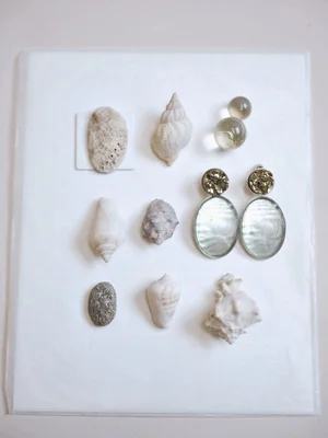 Still life arrangement of assorted seashells, a small smooth stone, glass spheres, and metallic drop earrings on white paper.