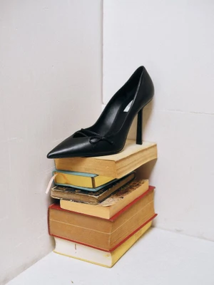 A black pointed-toe stiletto high heel resting atop a stack of vintage hardcover books against a white backdrop.