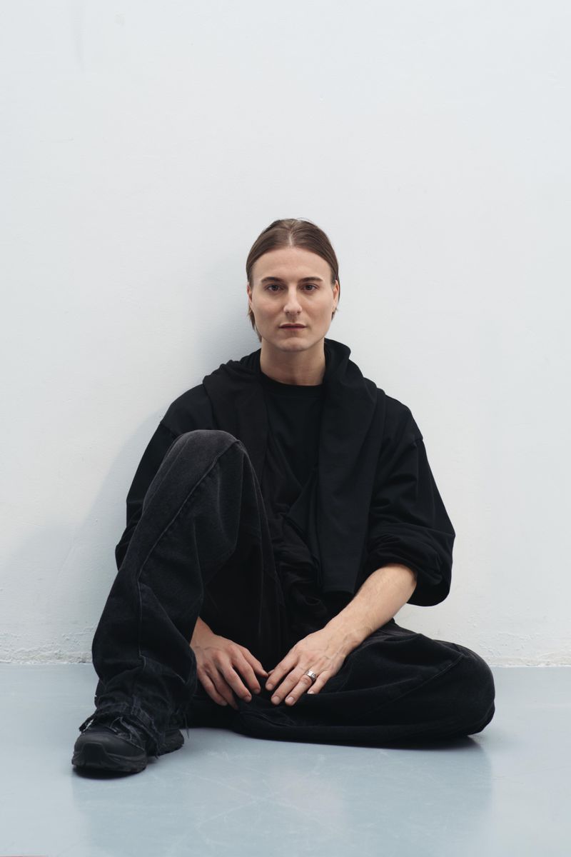 Person in an all-black layered outfit seated cross-legged against a stark white studio wall with hands resting on their lap.