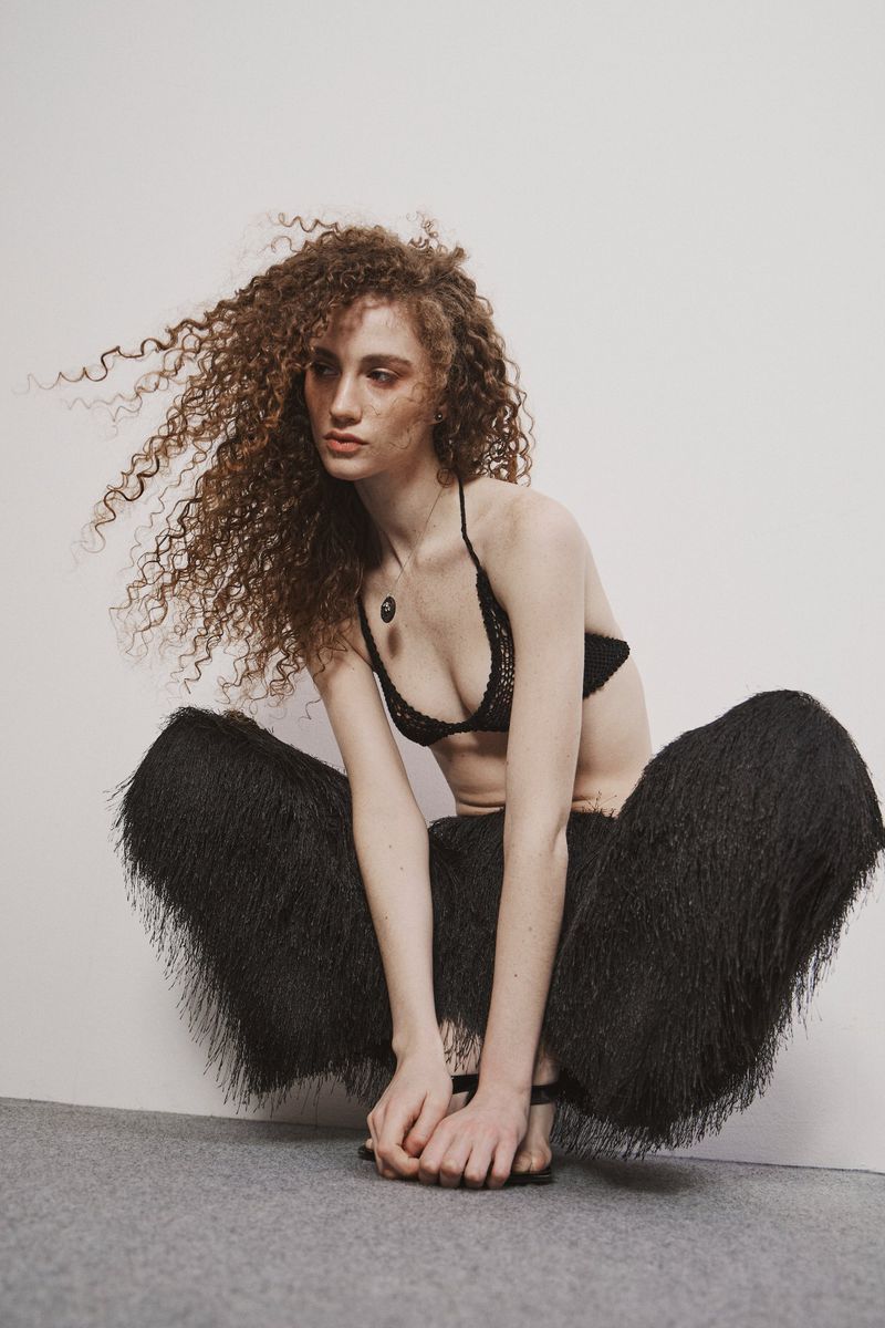 Model Roberta Laganà crouching in a black bralette and voluminous black fringe pants against a studio backdrop.