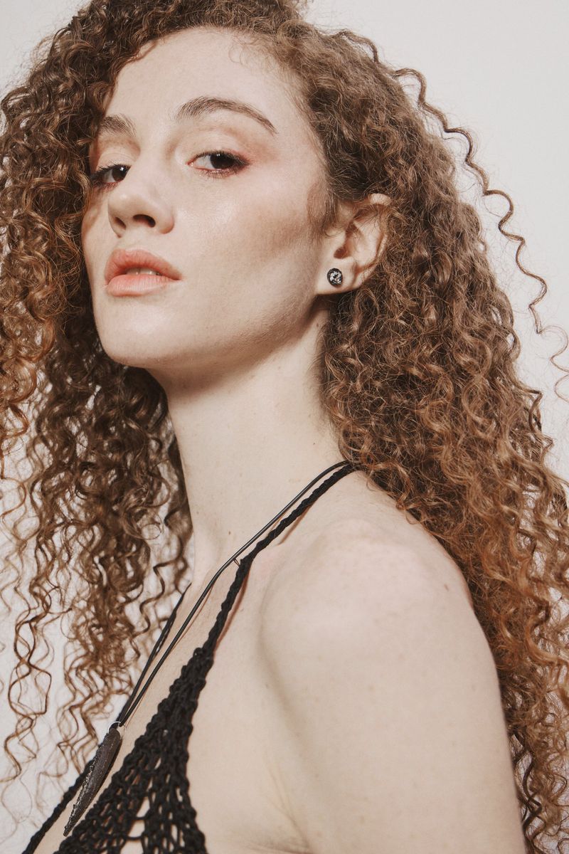 A woman with voluminous long curly brown hair wearing small stud earrings and a black knit halter top against a white studio backdrop.