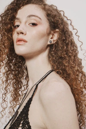 A woman with voluminous long curly brown hair wearing small stud earrings and a black knit halter top against a white studio backdrop.