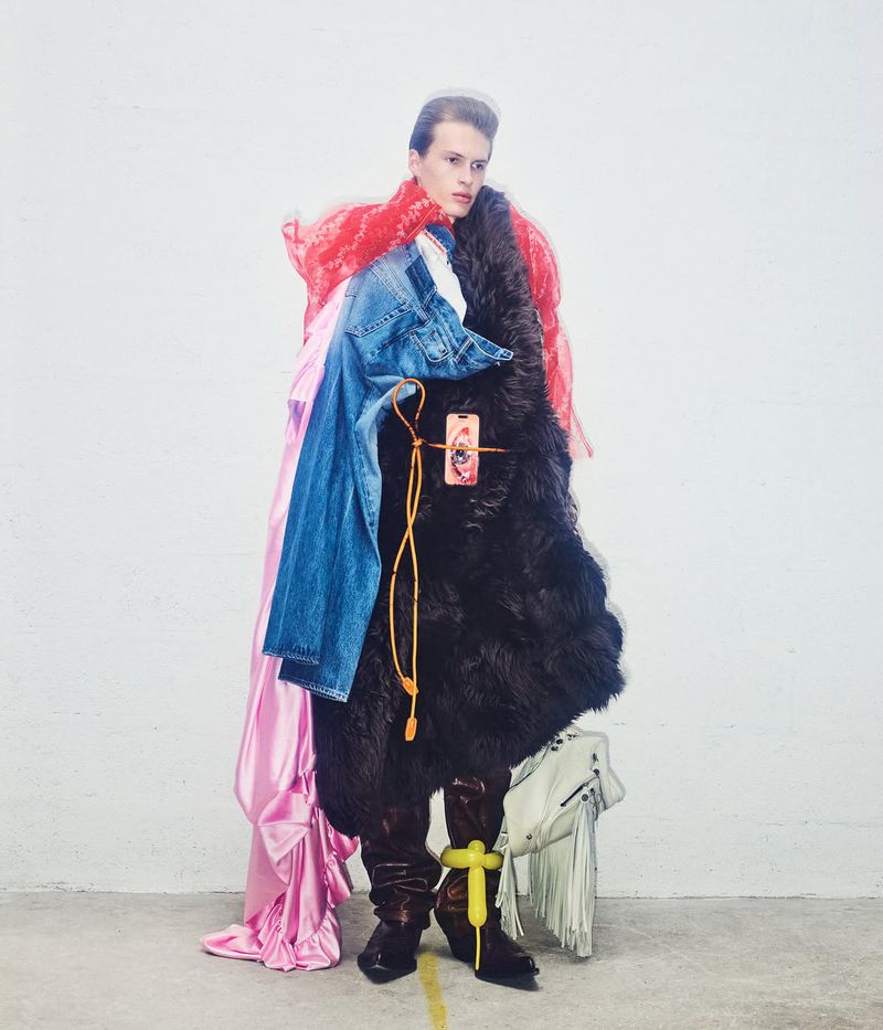 Full-length fashion portrait of a model draped in an eclectic mix of black fur, denim, and pink satin against a plain studio wall.