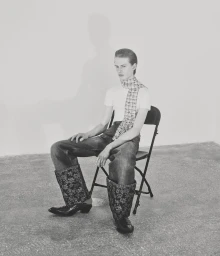 A man in a white t-shirt, patterned neck scarf, and jeans with wide floral cuffs sits on a metal folding chair in black and white.