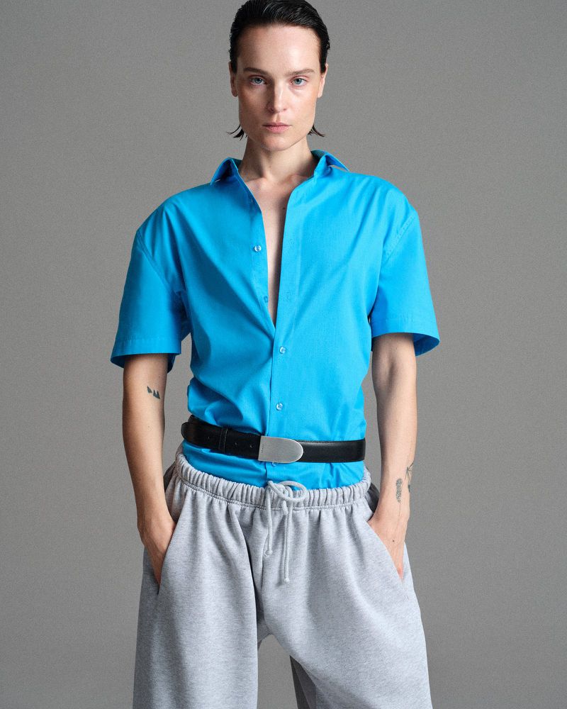 Model wearing a bright blue short-sleeve shirt belted over grey sweatpants against a neutral studio backdrop.