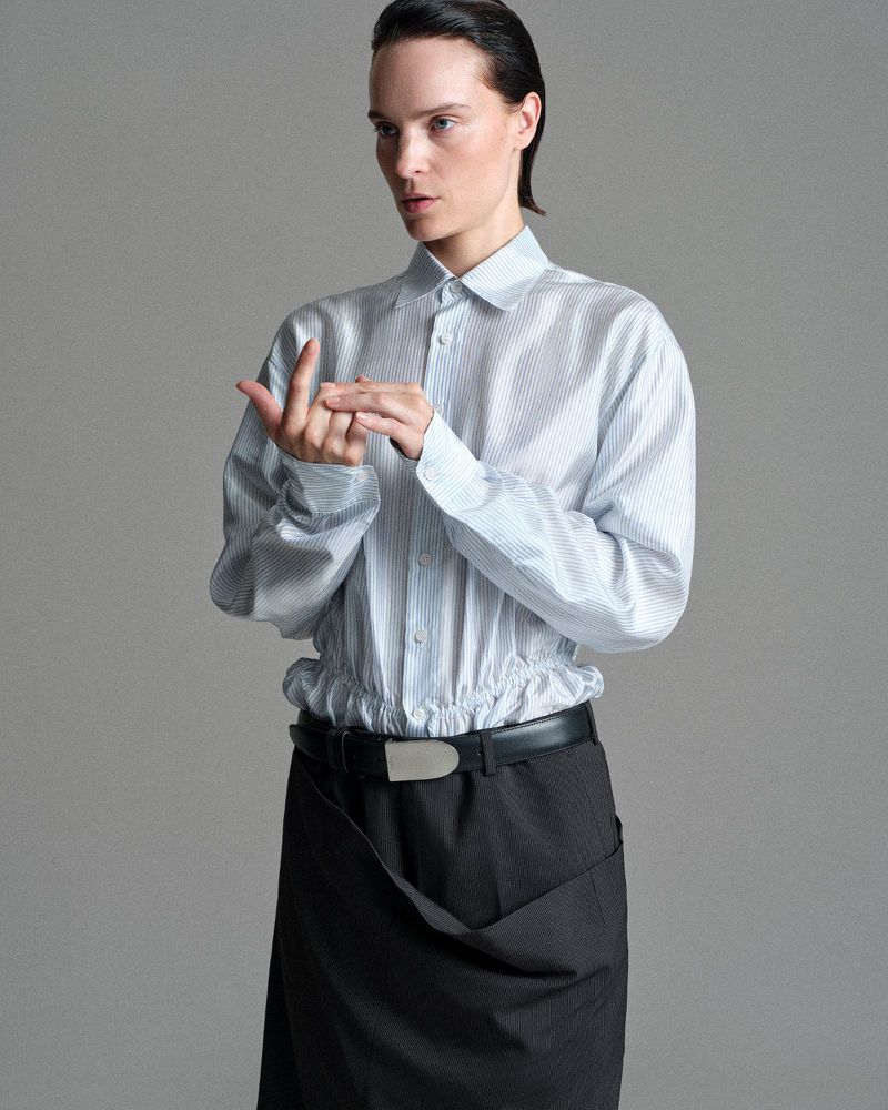 Model Charlotte Tomas poses in a striped button-up shirt and draped dark skirt with a wide belt against a grey studio backdrop.