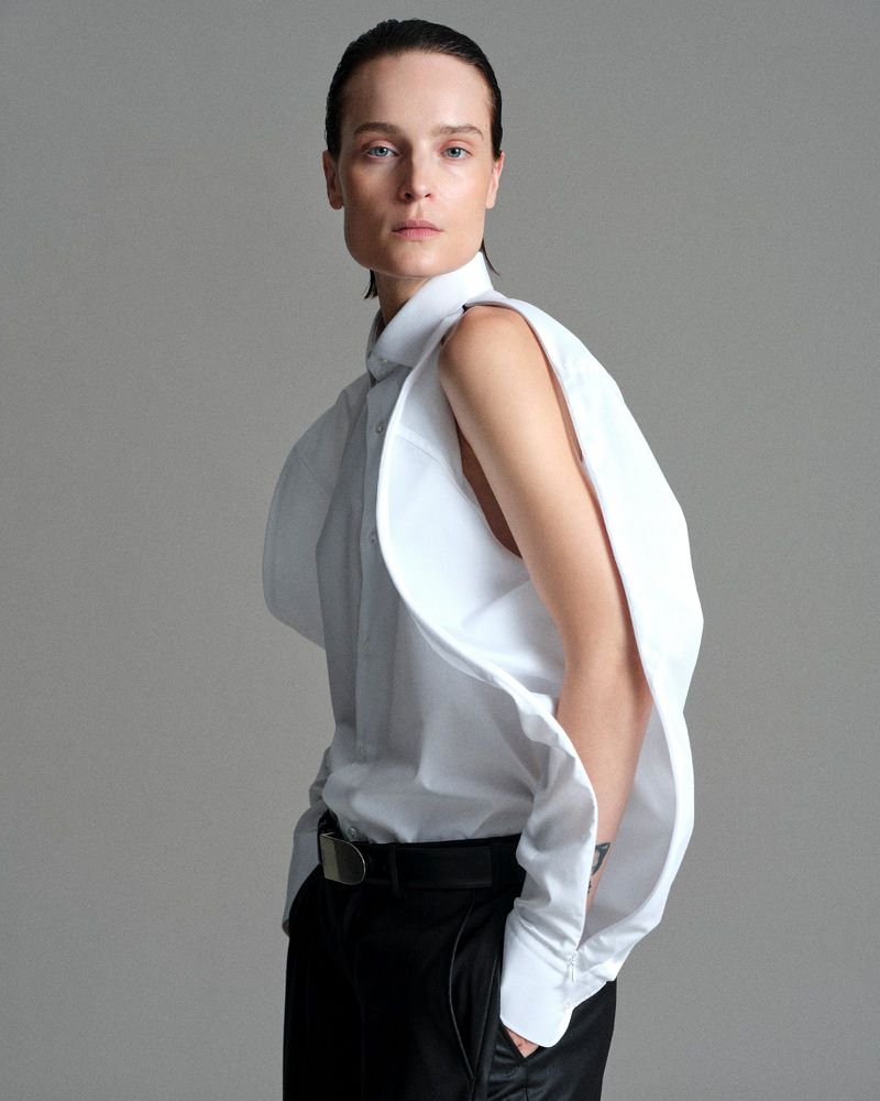 Model Charlotte Tomas wearing a deconstructed white cutout shirt and black trousers against a neutral grey studio background.
