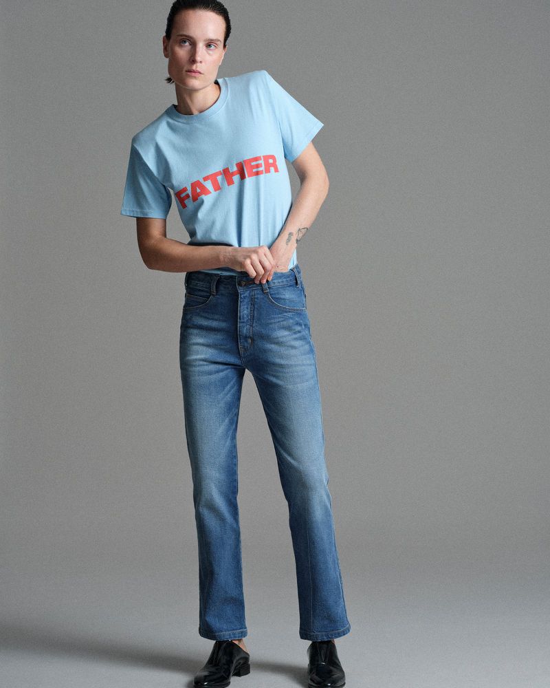Model Charlotte Tomas wearing a light blue FATHER graphic t-shirt and blue straight-leg denim jeans against a grey studio backdrop.