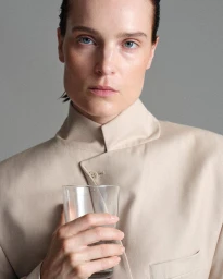 Charlotte Tomas wearing a beige high-collar tailored jacket and holding a clear glass of water against a grey studio backdrop.