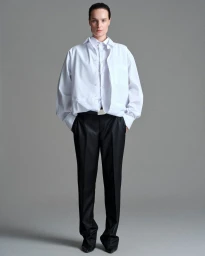Model Charlotte Tomas standing in an oversized layered white button-up shirt and tailored black trousers against a neutral grey studio backdrop.