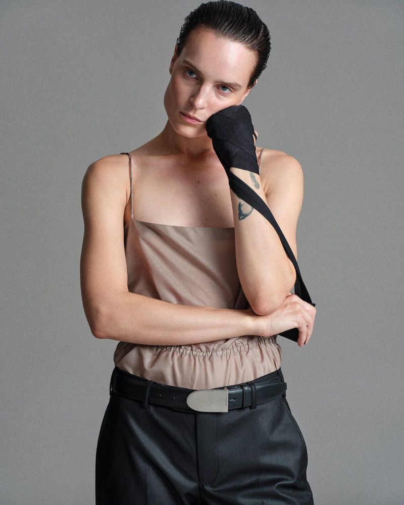 Model Charlotte Tomas in a taupe camisole and black trousers with a black hand wrap against a grey studio backdrop.