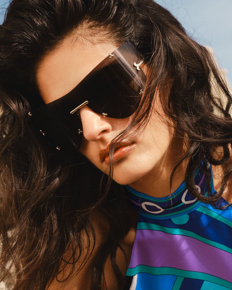 Close-up portrait of model Avishag Amir wearing oversized black shield sunglasses and a blue printed halter top.