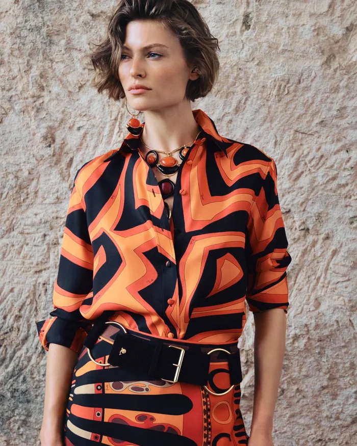 Model wearing a vibrant orange and black geometric patterned silk shirt and matching skirt against a textured stone wall.