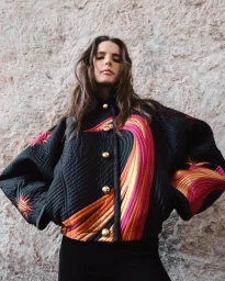 Model wearing a black quilted Emilio Pucci bomber jacket with colorful swirl accents and gold dome buttons against a stone wall.