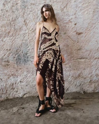 Model in an embellished asymmetrical black slip dress and ankle-tie sandals posing in front of a textured stone wall.